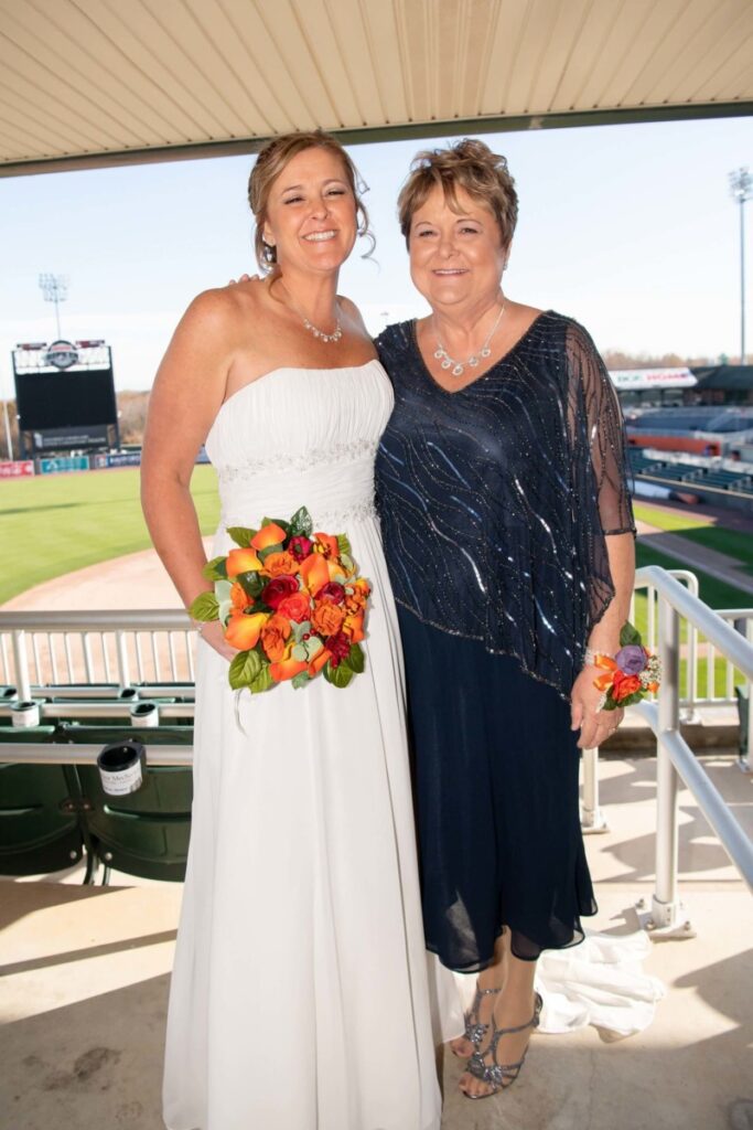 Heather-Scott-Wedding-Low-Res-19 1 - TOMMY GATZ ENTERTAINMENT Two women smiling; one in a white dress holding a bouquet, the other in a dark dress. They're standing in a stadium with empty seats and a baseball field in the background.