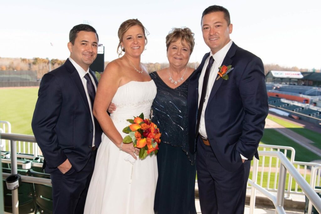Heather-Scott-Wedding-Low-Res-21 1 - TOMMY GATZ ENTERTAINMENT A bride in a white dress stands with two men in suits and a woman in a dark dress, holding a bouquet, with a sports field in the background.