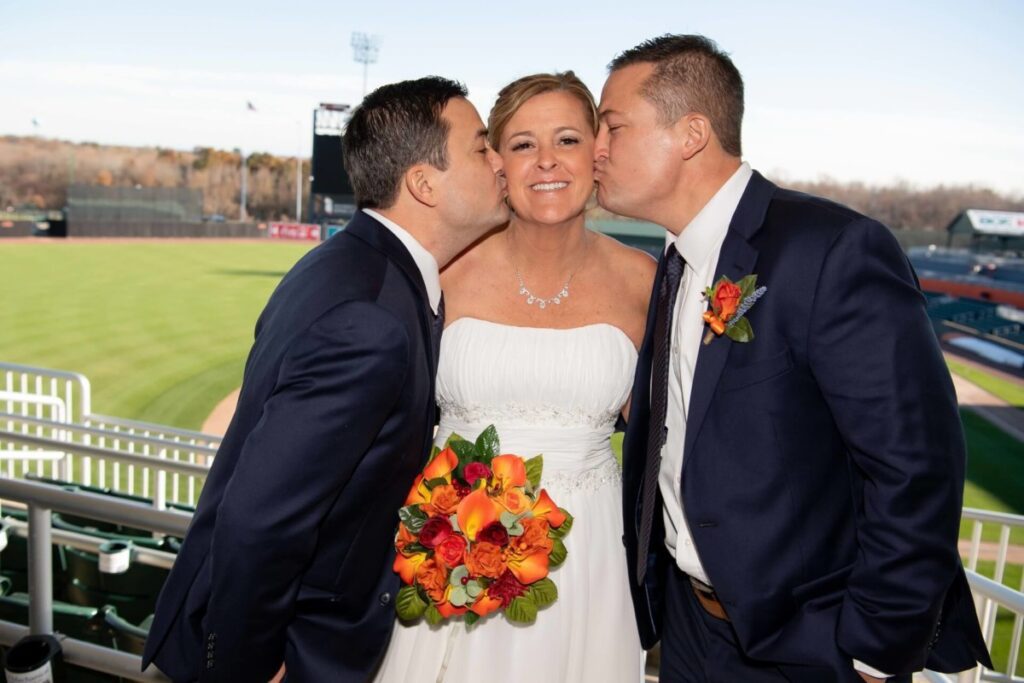 Heather-Scott-Wedding-Low-Res-22 1 - TOMMY GATZ ENTERTAINMENT Bride in a white dress holding flowers, standing between two men in suits kissing her cheeks, with a baseball field in the background.