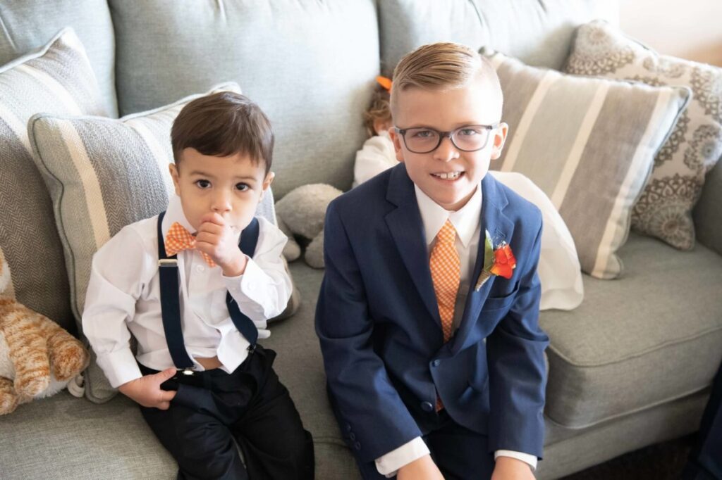 Heather-Scott-Wedding-Low-Res-23 1 - TOMMY GATZ ENTERTAINMENT Two young boys sit on a couch, both wearing white shirts and orange bow ties. One boy has suspenders; the other wears a suit jacket and glasses. A stuffed animal is beside them.