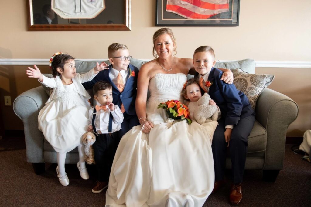 Heather-Scott-Wedding-Low-Res-24 1 - TOMMY GATZ ENTERTAINMENT A bride in a white gown sits on a couch with five children dressed in formal attire, smiling for the camera.