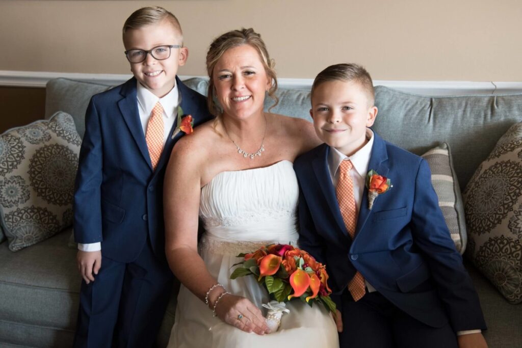Heather-Scott-Wedding-Low-Res-25 1 - TOMMY GATZ ENTERTAINMENT A smiling woman in a white dress sits on a couch between two young boys in blue suits with orange ties and boutonnieres.