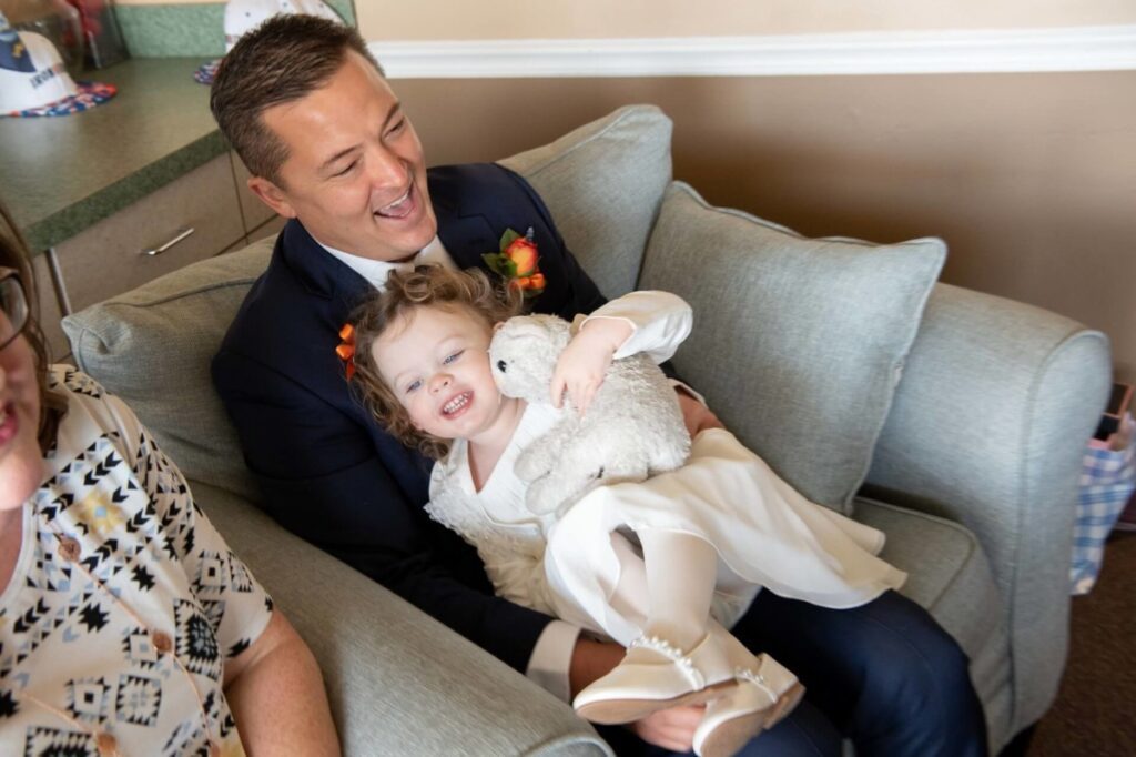 Heather-Scott-Wedding-Low-Res-26 1 - TOMMY GATZ ENTERTAINMENT A man in a suit sits on a sofa with a smiling child in a white dress holding a stuffed animal.