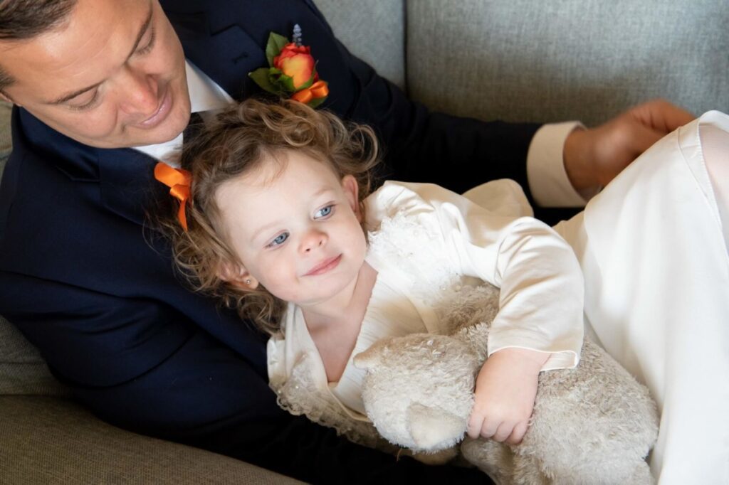Heather-Scott-Wedding-Low-Res-27 1 - TOMMY GATZ ENTERTAINMENT A man in a suit sits on a couch with a young child wearing a white dress, holding a stuffed animal. Flowers on the man's lapel suggest a formal occasion.