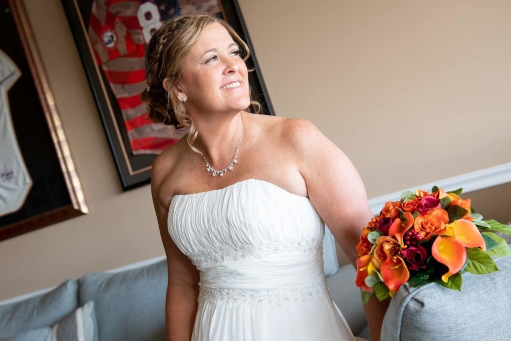 Heather-Scott-Wedding-Low-Res-28 1 - TOMMY GATZ ENTERTAINMENT A bride in a strapless white dress holds a vibrant bouquet of orange flowers while looking to the side, standing indoors with framed sports jerseys on the wall.
