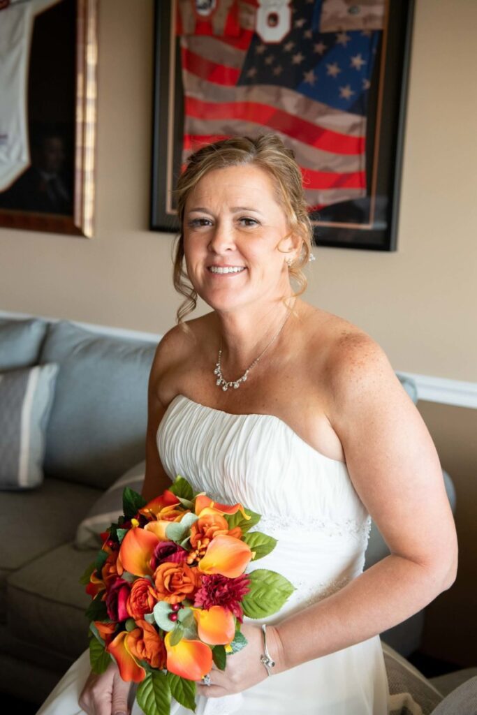 Heather-Scott-Wedding-Low-Res-29 1 - TOMMY GATZ ENTERTAINMENT A woman in a white dress holds a bouquet of vibrant orange and red flowers, sitting on a sofa with sports memorabilia in the background.