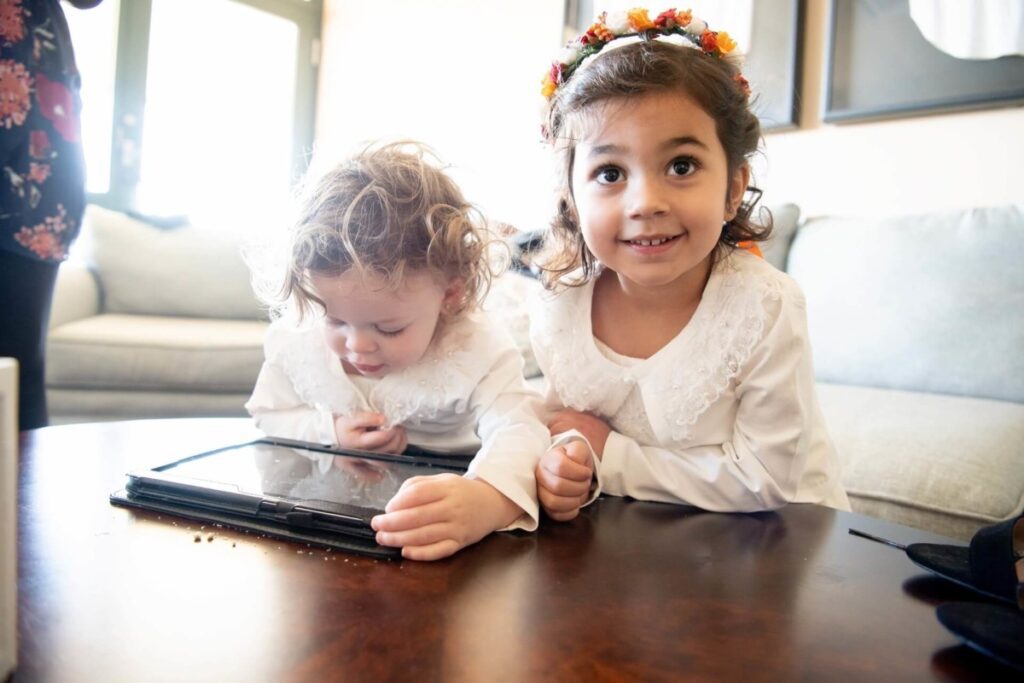 Heather-Scott-Wedding-Low-Res-3 1 - TOMMY GATZ ENTERTAINMENT Two young children in white shirts are engaged with a tablet on a wooden table. The older child smiles while the younger looks at the screen.