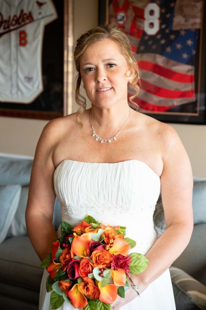 Heather-Scott-Wedding-Low-Res-30 1 - TOMMY GATZ ENTERTAINMENT A bride in a white dress holds a bouquet of orange and red flowers, standing in front of framed sports jerseys and an American flag.
