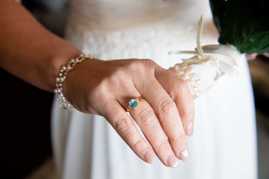 Heather-Scott-Wedding-Low-Res-32 1 - TOMMY GATZ ENTERTAINMENT Close-up of a hand showing an engagement ring with a turquoise gemstone. The hand rests on a lacy fabric and wears a pearl bracelet.