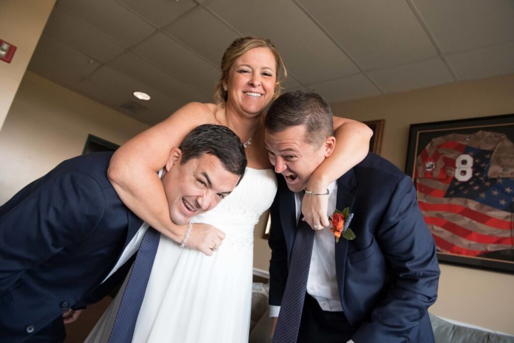 Heather-Scott-Wedding-Low-Res-33 1 - TOMMY GATZ ENTERTAINMENT A woman in a white dress playfully wraps her arms around two men in suits. They are all smiling and appear to be having fun indoors.