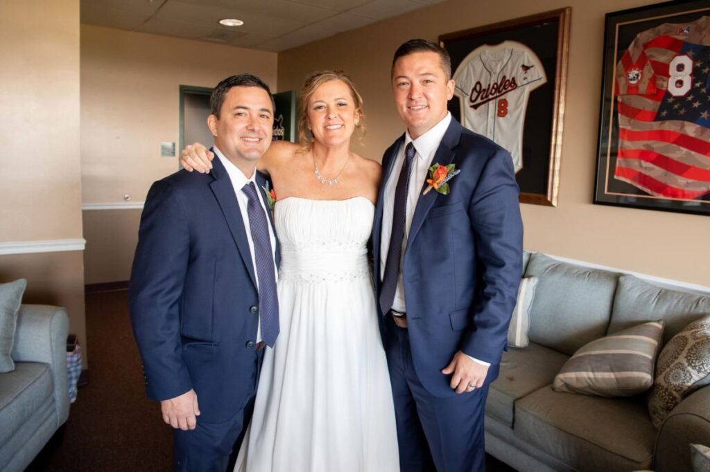 Heather-Scott-Wedding-Low-Res-34 1 - TOMMY GATZ ENTERTAINMENT Three people dressed in formal attire pose together in a room with sports memorabilia on the walls.