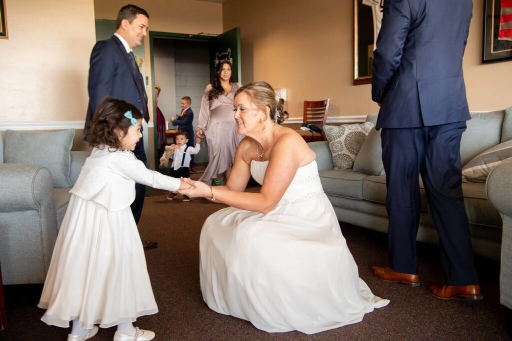 Heather-Scott-Wedding-Low-Res-35 1 - TOMMY GATZ ENTERTAINMENT A bride in a white dress kneels to greet a young girl in a white outfit. Several adults and children are in the room, with some standing and some seated.