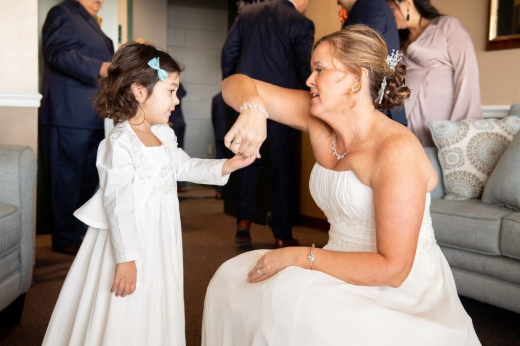 Heather-Scott-Wedding-Low-Res-36 1 - TOMMY GATZ ENTERTAINMENT A woman in a white dress greets a young girl in a matching dress. They hold hands in a living room with people in the background.