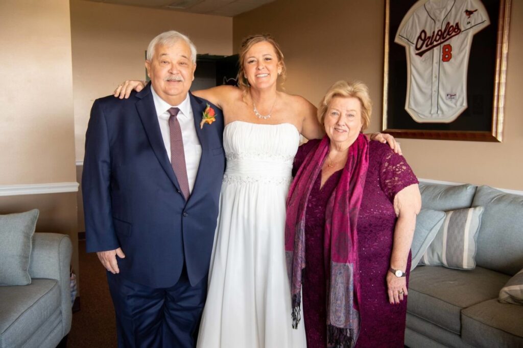Heather-Scott-Wedding-Low-Res-37 1 - TOMMY GATZ ENTERTAINMENT Three people pose together in a room; a woman in a white dress stands between a man in a suit and a woman in a purple dress. An Orioles jersey is framed on the wall behind them.