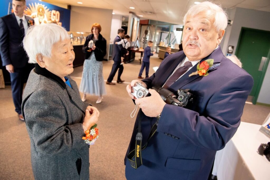 Heather-Scott-Wedding-Low-Res-39 1 - TOMMY GATZ ENTERTAINMENT Two people in formal attire engage in conversation, both holding cameras. Others in the background carry drinks and chat in a brightly lit room.