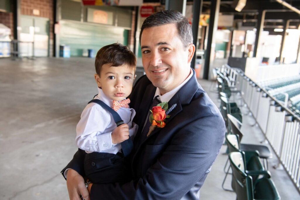 Heather-Scott-Wedding-Low-Res-40 1 - TOMMY GATZ ENTERTAINMENT A man in a suit holds a young child wearing a bow tie in an indoor setting with seating and overhead lights.