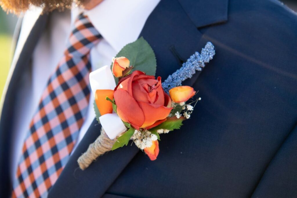 Heather-Scott-Wedding-Low-Res-41 1 - TOMMY GATZ ENTERTAINMENT A close-up of a man's suit jacket with a boutonniere featuring an orange rose, small buds, and a sprig of lavender. The tie has an orange and navy plaid pattern.