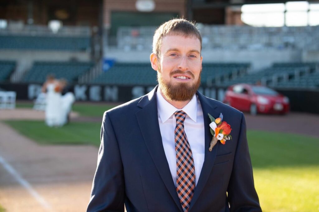 Heather-Scott-Wedding-Low-Res-42 1 - TOMMY GATZ ENTERTAINMENT Man in a suit with a checked tie and boutonniere stands on a sports field, with a red car and seating area in the background.