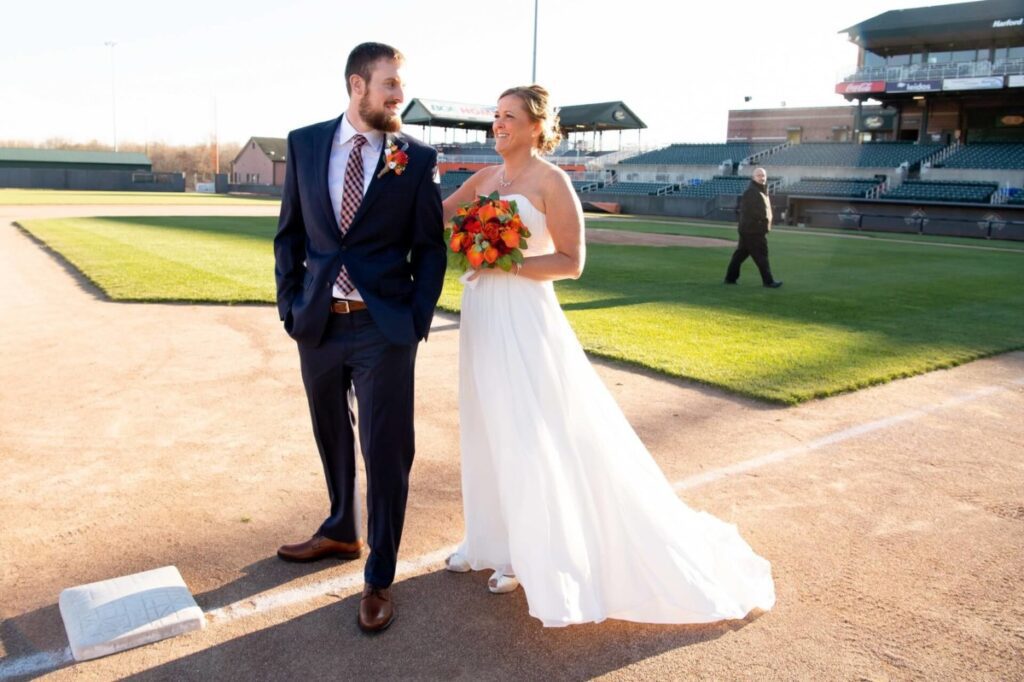 Heather-Scott-Wedding-Low-Res-44 1 - TOMMY GATZ ENTERTAINMENT A bride and groom smile at each other while walking on a baseball field. The bride holds a bouquet of orange flowers.