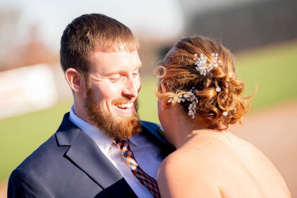 Heather-Scott-Wedding-Low-Res-46 1 - TOMMY GATZ ENTERTAINMENT A couple smiles together in an outdoor setting. The man has a beard and wears a suit, while the woman has styled hair with a jeweled accessory. The background is blurred.
