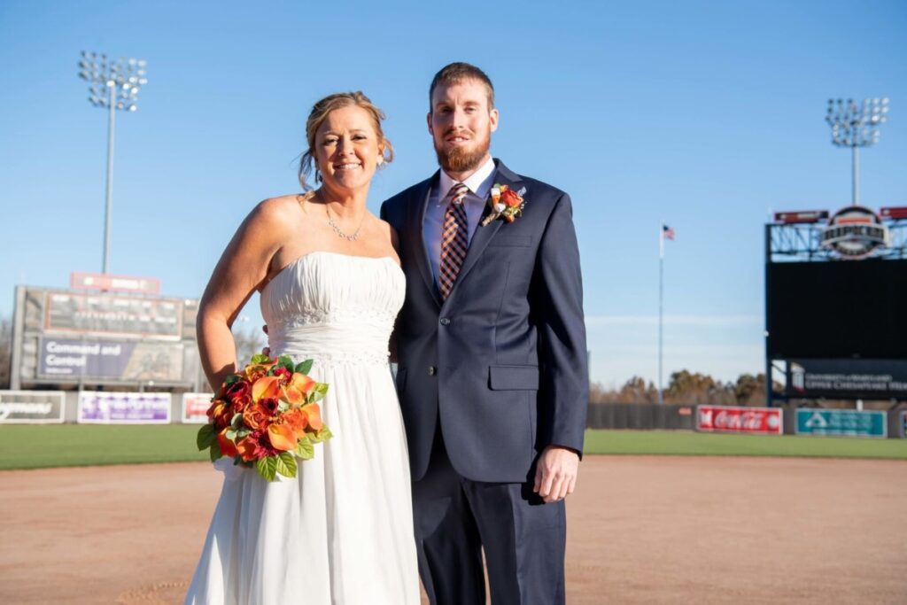 Heather-Scott-Wedding-Low-Res-47 1 - TOMMY GATZ ENTERTAINMENT A bride in a strapless white gown holds a bouquet while standing beside a groom in a navy suit with a striped tie on a baseball field.