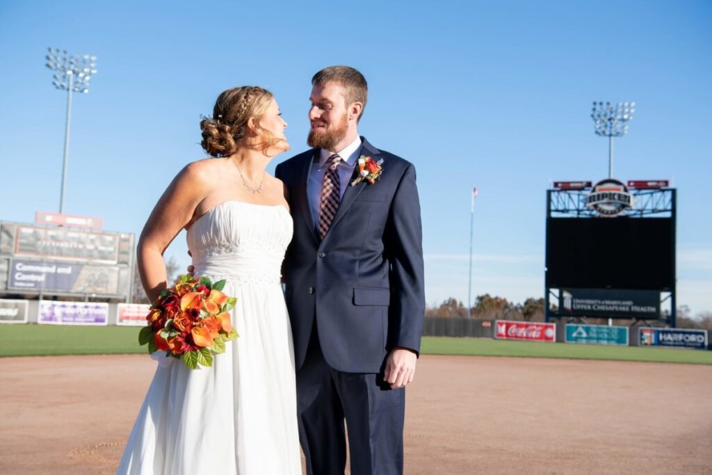 Heather-Scott-Wedding-Low-Res-48 1 - TOMMY GATZ ENTERTAINMENT A bride and groom stand on a baseball field. The bride holds a bouquet of orange flowers, and both are dressed in formal attire. The scoreboard and stands are visible in the background.
