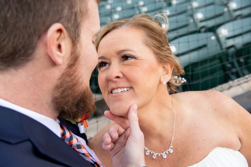 Heather-Scott-Wedding-Low-Res-49 1 - TOMMY GATZ ENTERTAINMENT A couple faces each other closely. The woman smiles gently, adorned with a jeweled necklace. The background shows stadium seating and a safety net.