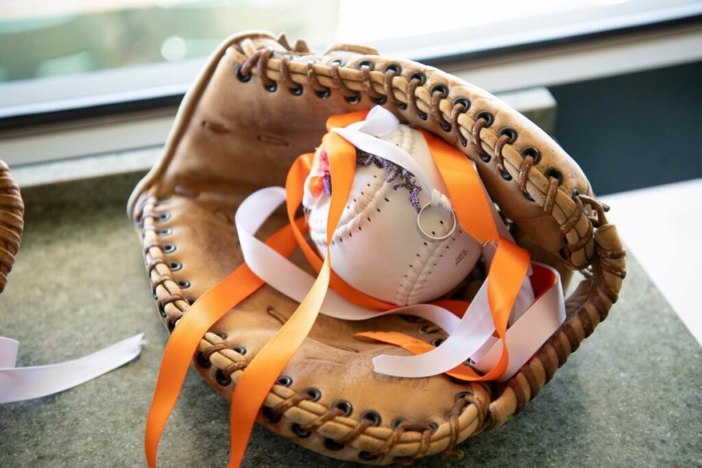 A baseball glove holds a baseball decorated with orange and white ribbons.