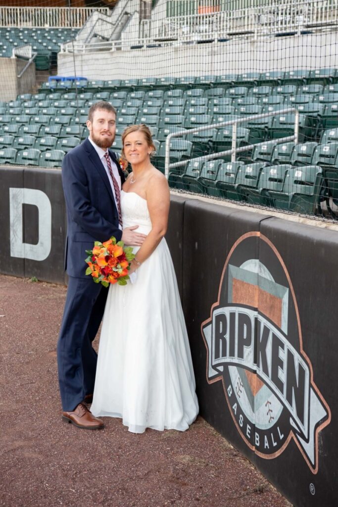 Heather-Scott-Wedding-Low-Res-50 1 - TOMMY GATZ ENTERTAINMENT A bride and groom stand by a "Ripken Baseball" sign in a baseball stadium. The bride holds a bouquet.