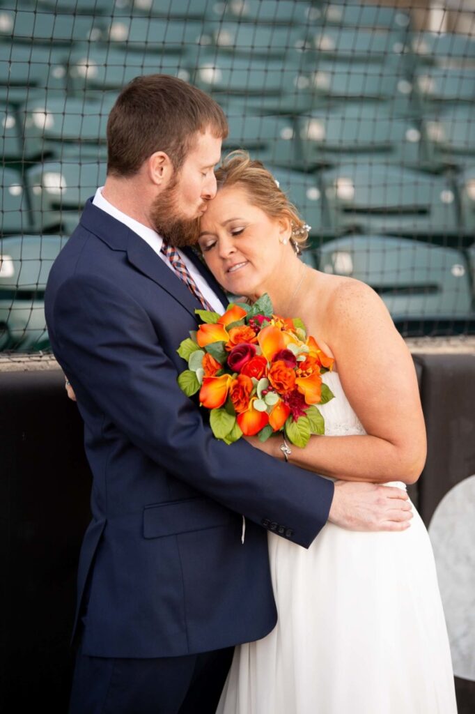 Heather-Scott-Wedding-Low-Res-51 1 - TOMMY GATZ ENTERTAINMENT A couple embraces, with the groom in a suit and the bride holding a bouquet of orange flowers. They are at a stadium near a netted barrier.