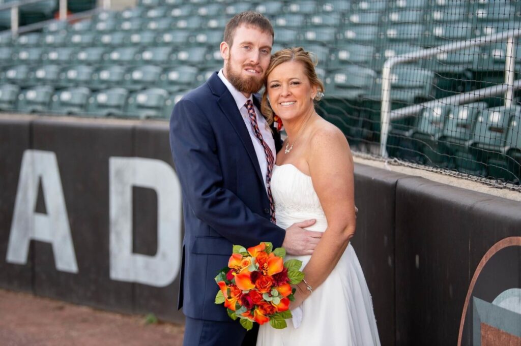 Heather-Scott-Wedding-Low-Res-52 1 - TOMMY GATZ ENTERTAINMENT A couple in wedding attire stands together on a baseball field, with the bride holding a bouquet of orange flowers.