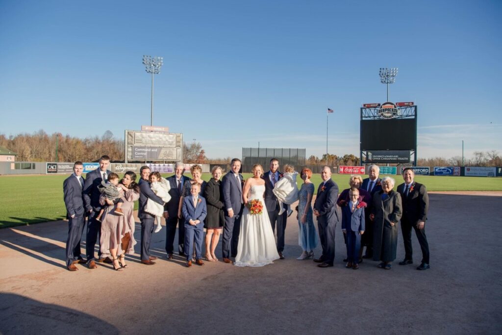 Heather-Scott-Wedding-Low-Res-53 1 - TOMMY GATZ ENTERTAINMENT A wedding party poses on a baseball field, with the bride and groom in the center, surrounded by family and friends under a clear blue sky.