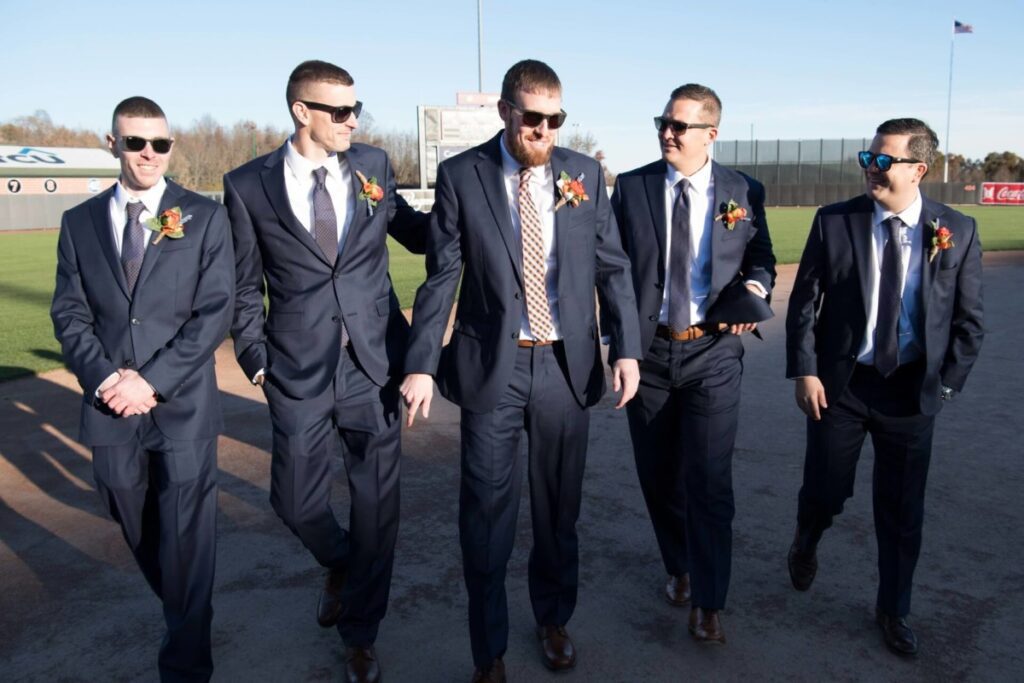 Heather-Scott-Wedding-Low-Res-54 1 - TOMMY GATZ ENTERTAINMENT Five men in suits and sunglasses walk together on a sports field.