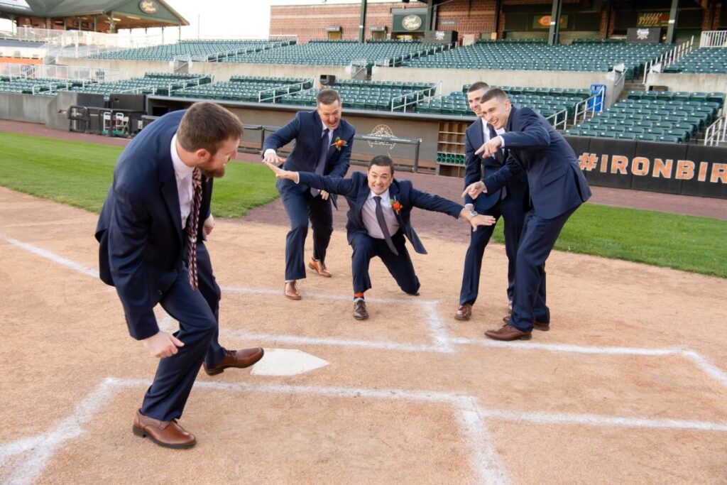 Heather-Scott-Wedding-Low-Res-55 1 - TOMMY GATZ ENTERTAINMENT A group of men in suits engage in playful poses on a baseball field's home plate.