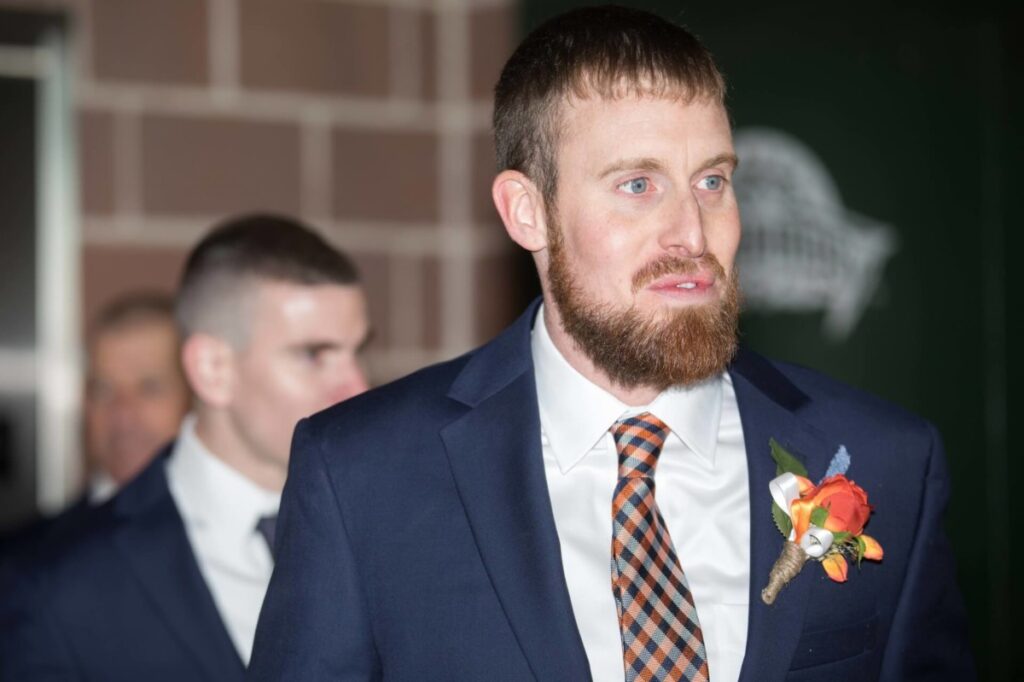 Heather-Scott-Wedding-Low-Res-58 1 - TOMMY GATZ ENTERTAINMENT Two men in suits, one with a beard and a patterned tie, with a floral boutonniere. The background is a brick wall and a dark green sign.