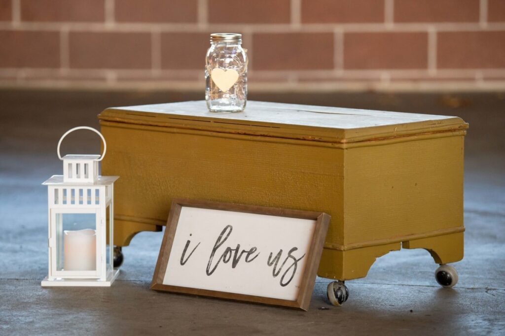 Heather-Scott-Wedding-Low-Res-59 1 - TOMMY GATZ ENTERTAINMENT A yellow trunk with a jar and a white lantern on top is on a concrete floor. A framed sign leaning against it reads "i love us.