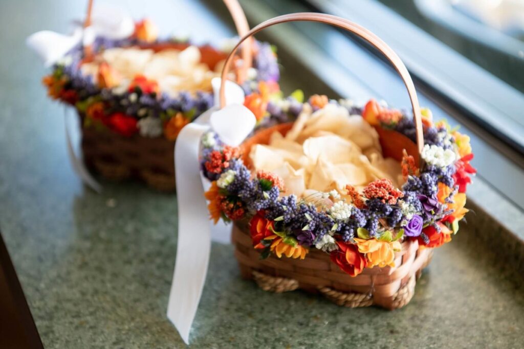 Heather-Scott-Wedding-Low-Res-6 1 - TOMMY GATZ ENTERTAINMENT Two wicker baskets adorned with colorful flowers and ribbons, filled with beige petals, placed on a green surface.
