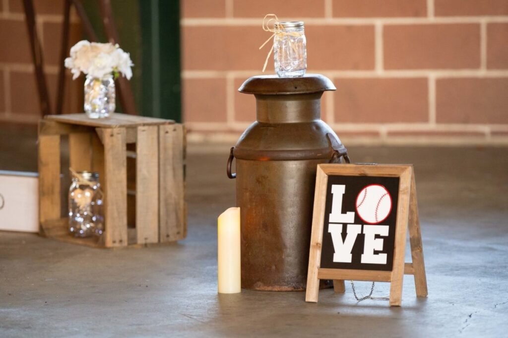 Heather-Scott-Wedding-Low-Res-60 1 - TOMMY GATZ ENTERTAINMENT A rustic setup with a metal milk can, a wooden crate with jars, a candle, and a small chalkboard sign displaying "LOVE" with a baseball replacing the "O".
