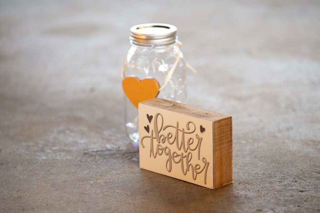 Heather-Scott-Wedding-Low-Res-61 1 - TOMMY GATZ ENTERTAINMENT A glass jar with a heart and a wooden block displaying the text "better together" are placed on a smooth gray surface.
