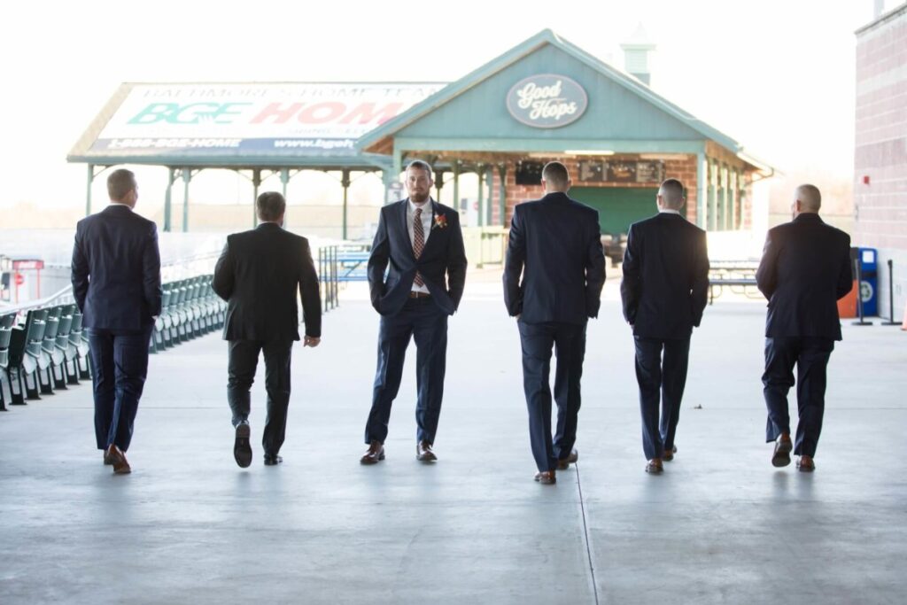 Heather-Scott-Wedding-Low-Res-62 1 - TOMMY GATZ ENTERTAINMENT Six men in suits walk away from the camera in an outdoor setting, with a pavilion in the background.