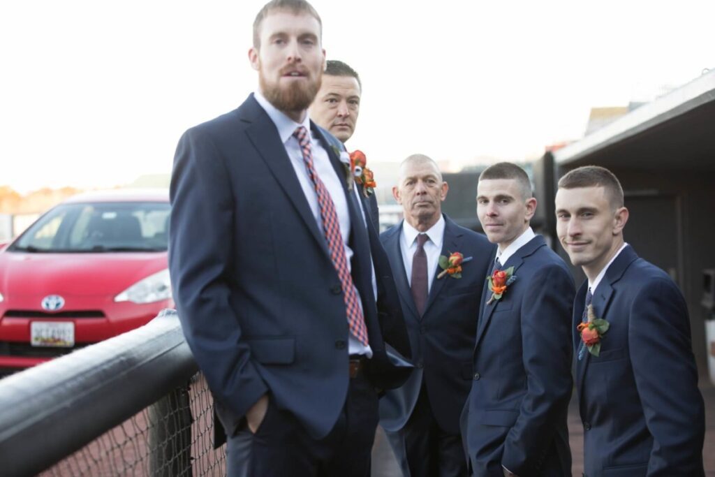 Heather-Scott-Wedding-Low-Res-63 1 - TOMMY GATZ ENTERTAINMENT Five men in suits with red and orange boutonnieres stand outside near a red car.