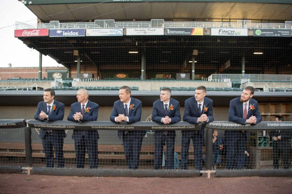 Heather-Scott-Wedding-Low-Res-64 1 - TOMMY GATZ ENTERTAINMENT Six men in suits stand behind a baseball dugout fence, wearing boutonnieres.
