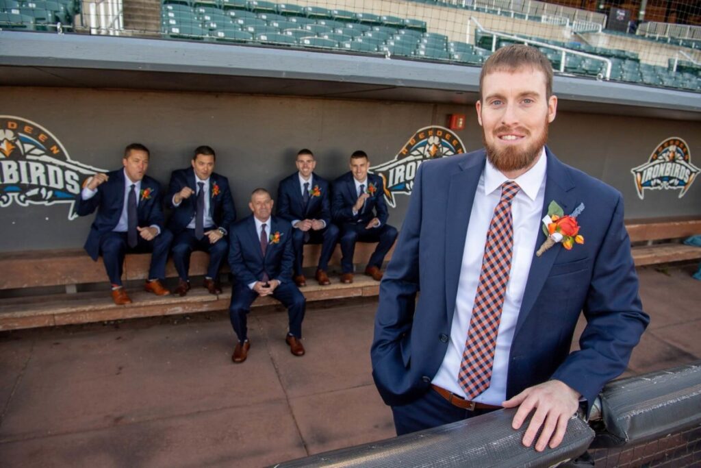 Heather-Scott-Wedding-Low-Res-65 1 - TOMMY GATZ ENTERTAINMENT A man in a suit with a flower boutonniere stands in the foreground, while five other men in suits are seated in the background of a dugout with baseball team logos.