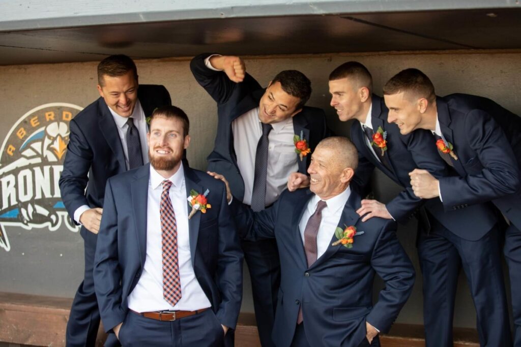 Heather-Scott-Wedding-Low-Res-66 1 - TOMMY GATZ ENTERTAINMENT Six men in suits, with colorful boutonnières, pose playfully in a dugout.
