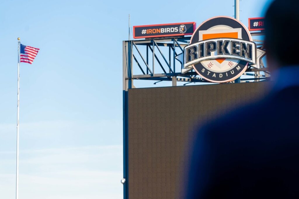 Heather-Scott-Wedding-Low-Res-67 1 - TOMMY GATZ ENTERTAINMENT Ripken Stadium sign with #IronBirds hashtag against a clear blue sky and an American flag on a pole nearby. Large dark silhouette in the foreground.