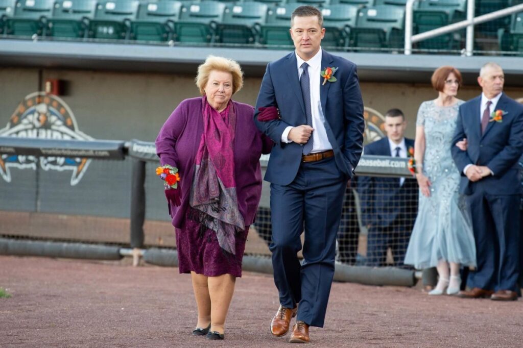 Heather-Scott-Wedding-Low-Res-69 1 - TOMMY GATZ ENTERTAINMENT A woman in a purple outfit walks arm-in-arm with a man in a blue suit on a baseball field. Others in formal attire are in the background.