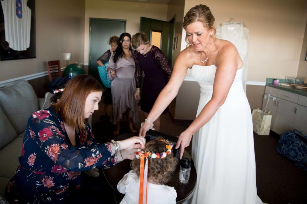Heather-Scott-Wedding-Low-Res-7 1 - TOMMY GATZ ENTERTAINMENT A bride in a white dress adjusts a child's hair with help from another woman in a floral blouse. Several people are in the background in a room.