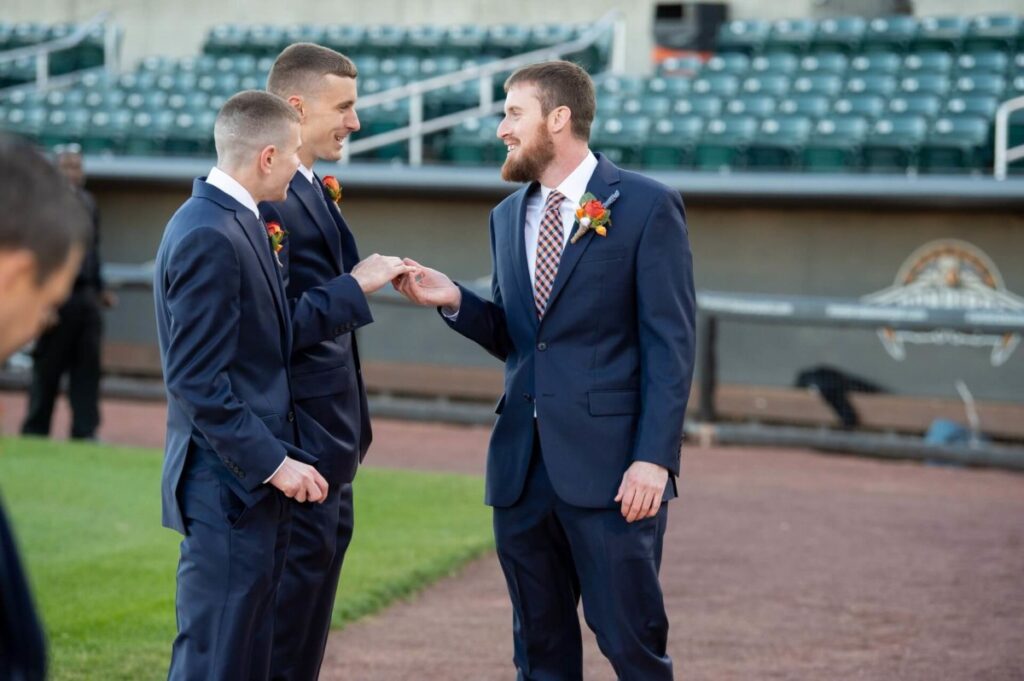Heather-Scott-Wedding-Low-Res-70 1 - TOMMY GATZ ENTERTAINMENT Three men in suits converse and smile on a baseball field.