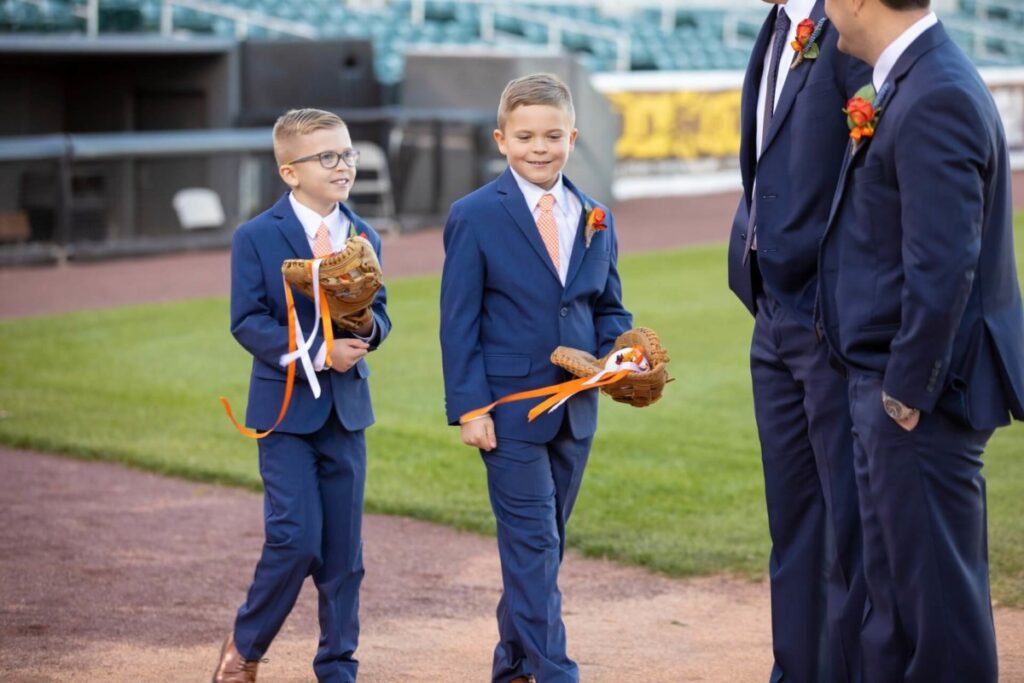 Heather-Scott-Wedding-Low-Res-71 2 - TOMMY GATZ ENTERTAINMENT Two boys in blue suits walk on a baseball field holding gloves with orange ribbons. Two adults in similar attire walk beside them.