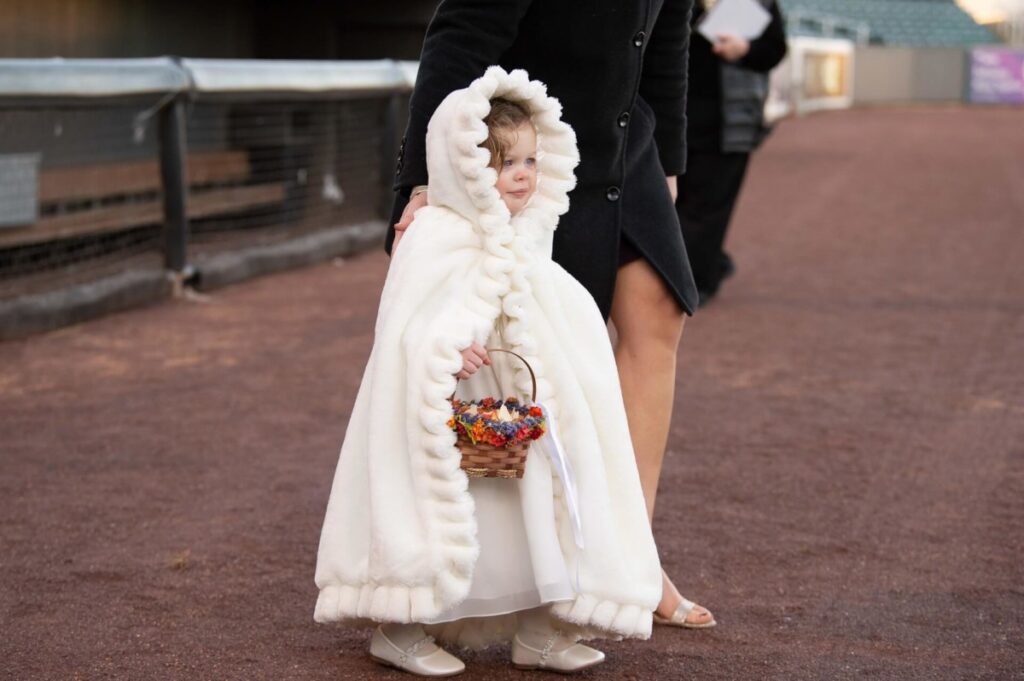 Heather-Scott-Wedding-Low-Res-72 1 - TOMMY GATZ ENTERTAINMENT A child in a white, hooded cloak holds a basket of flowers. They stand on a path next to an adult in a dark coat.